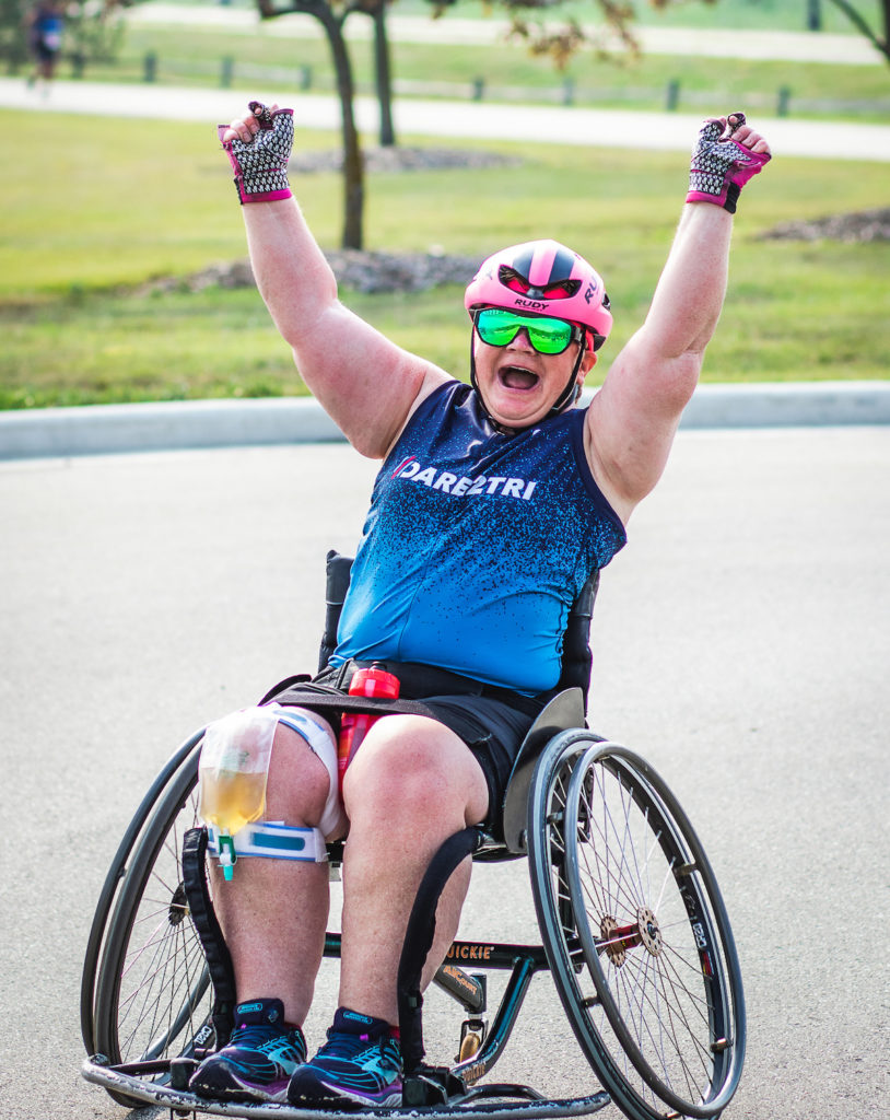 Kelly Butler sitting in a sports wheelchair. She's wearing a Dare2tri triathlon top, smiling with her arms raised.