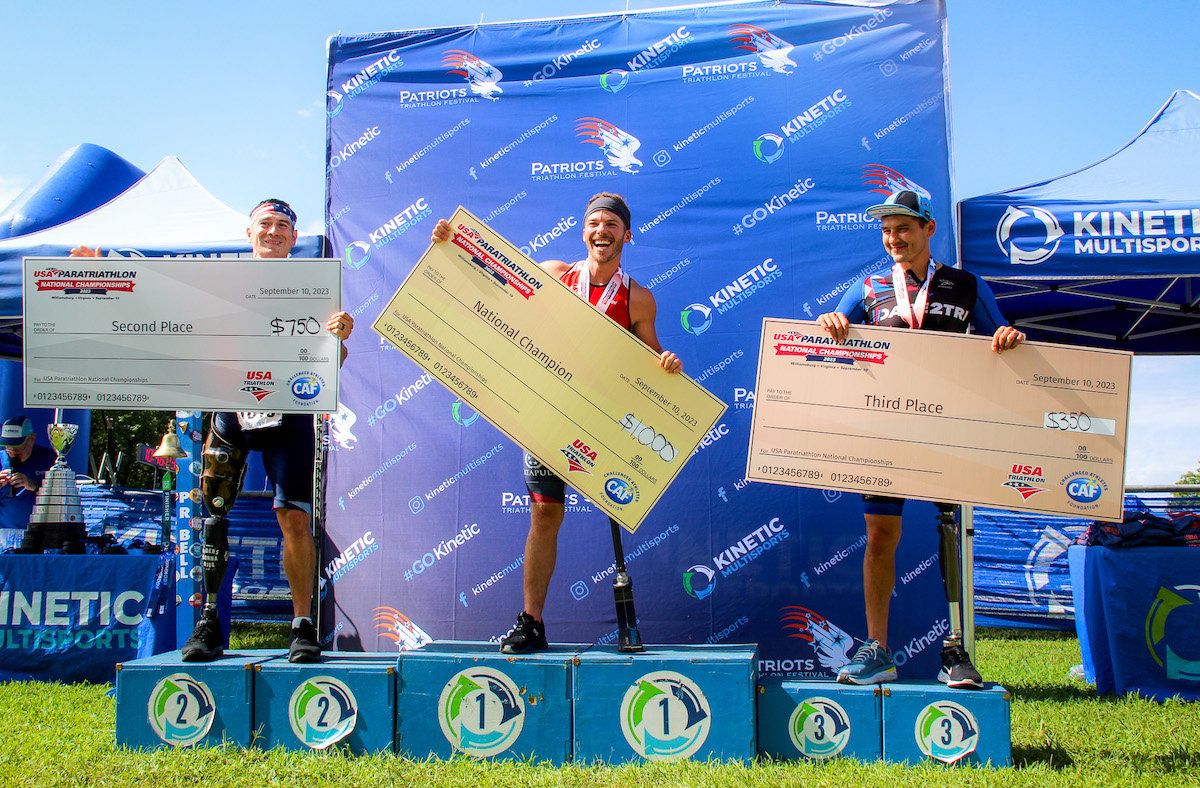 Three men standing atop a race podium holding giant checks.