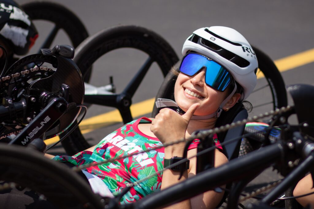 A woman in a recumbent handcycle smiling and giving a thumbs up.